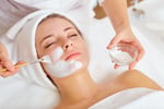 A young woman with a towel wrapped around her head receives a relaxing facial treatment. A spa professional is applying a white facial mask with a brush while holding a small glass bowl of cream. The woman appears calm and serene.