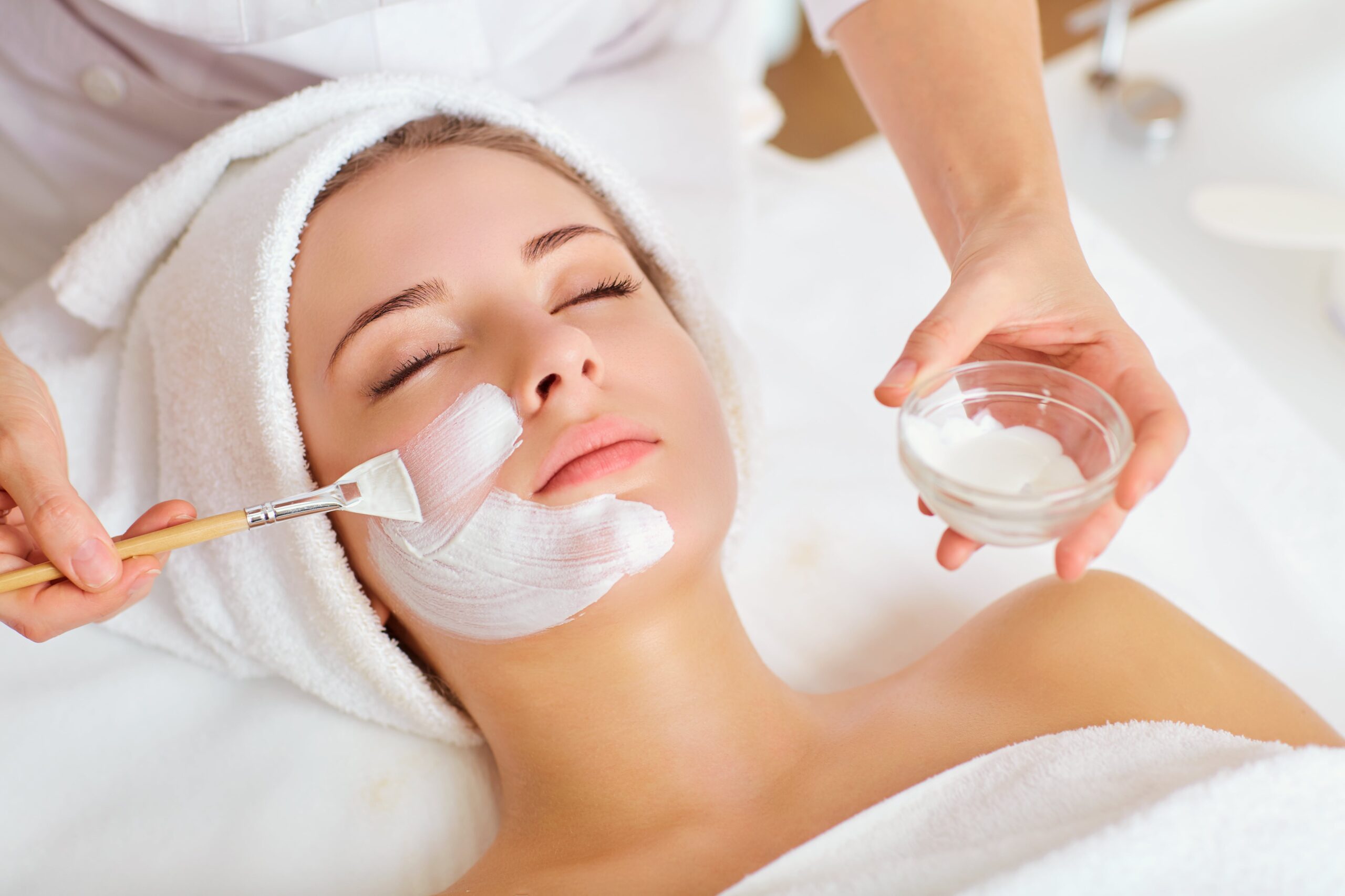 Facial Spa Treatment A young woman with a towel wrapped around her head receives a relaxing facial treatment. A spa professional is applying a white facial mask with a brush while holding a small glass bowl of cream. The woman appears calm and serene.